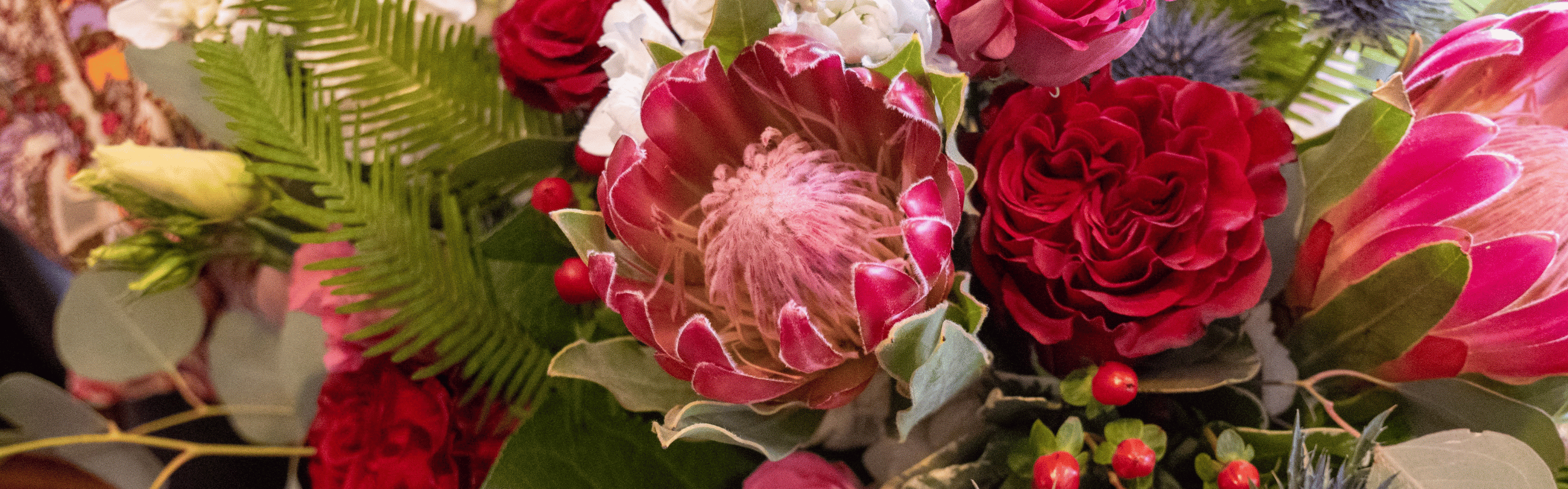 Vibrant floral arrangement with pink King Protea, red roses, white flowers, green ferns, and red berries.